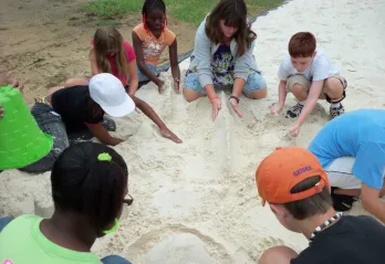 A group of children sit in a circle with their group leader, working together on structures in a sandbox.