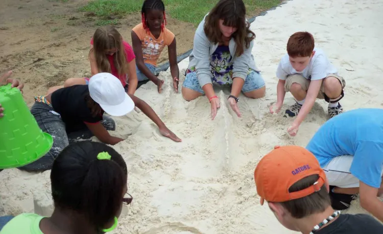 A group of children sit in a circle with their group leader, working together on structures in a sandbox.