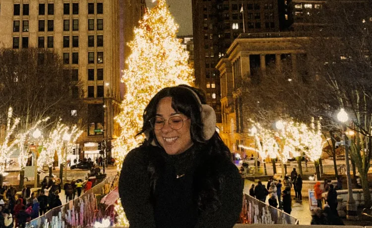 Photo of engagement staff member Adriana, posing for a photo in a park with a Christmas tree and lights in the background.